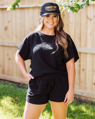 Full-body shot of a woman in a black two-piece casual outfit with a 'Cowboy Hat' trucker cap