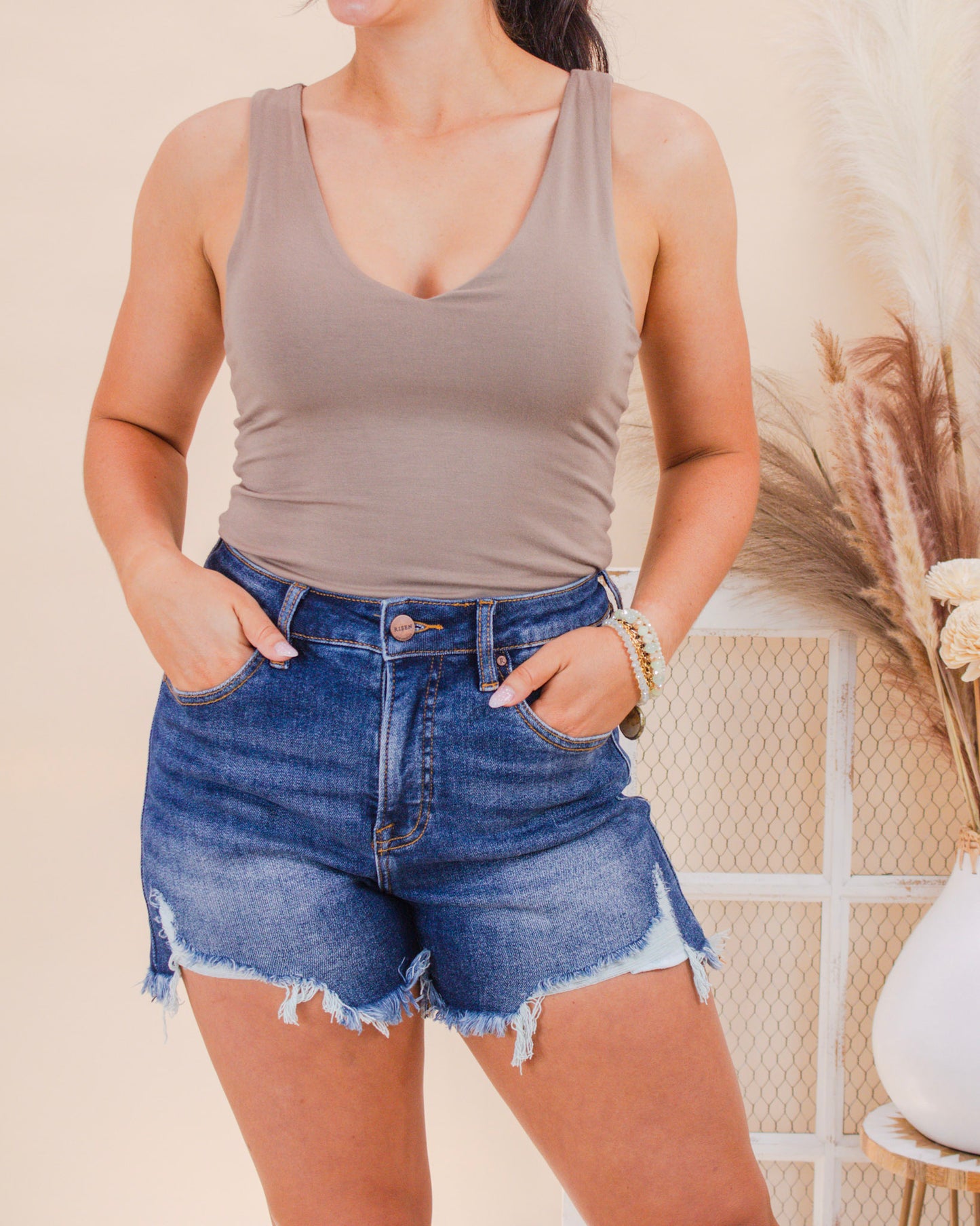 A woman models a neutral taupe tank top paired with high-waisted distressed denim shorts, styled for a casual summer look with layered bracelets and natural textures.