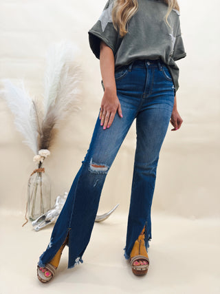 Woman wearing distressed high-rise flare jeans with front ankle slits and raw hems, styled with chunky platform sandals and an oversized olive graphic tee.