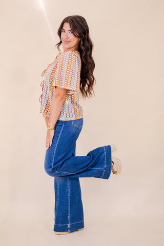 Woman wearing a colorful crochet-style tie-front top with flutter sleeves, paired with high-rise denim flare jeans and platform heels. A retro-inspired, boho-chic outfit perfect for casual summer days or festivals.