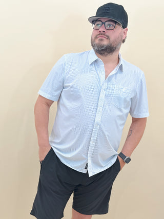 Men’s casual summer outfit featuring a white micro-print short sleeve button-down shirt by Flag & Anthem, paired with black shorts and a black cap.