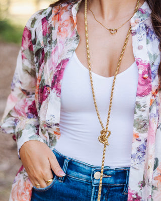 Close-up of a woman wearing a white tank top layered with a sheer floral button-down shirt, styled with gold layered necklaces and high-waisted denim jeans.