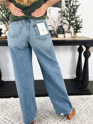 Back view of woman wearing high-rise, wide-leg blue jeans with raw hems and a visible brand tag in a cozy, styled room.