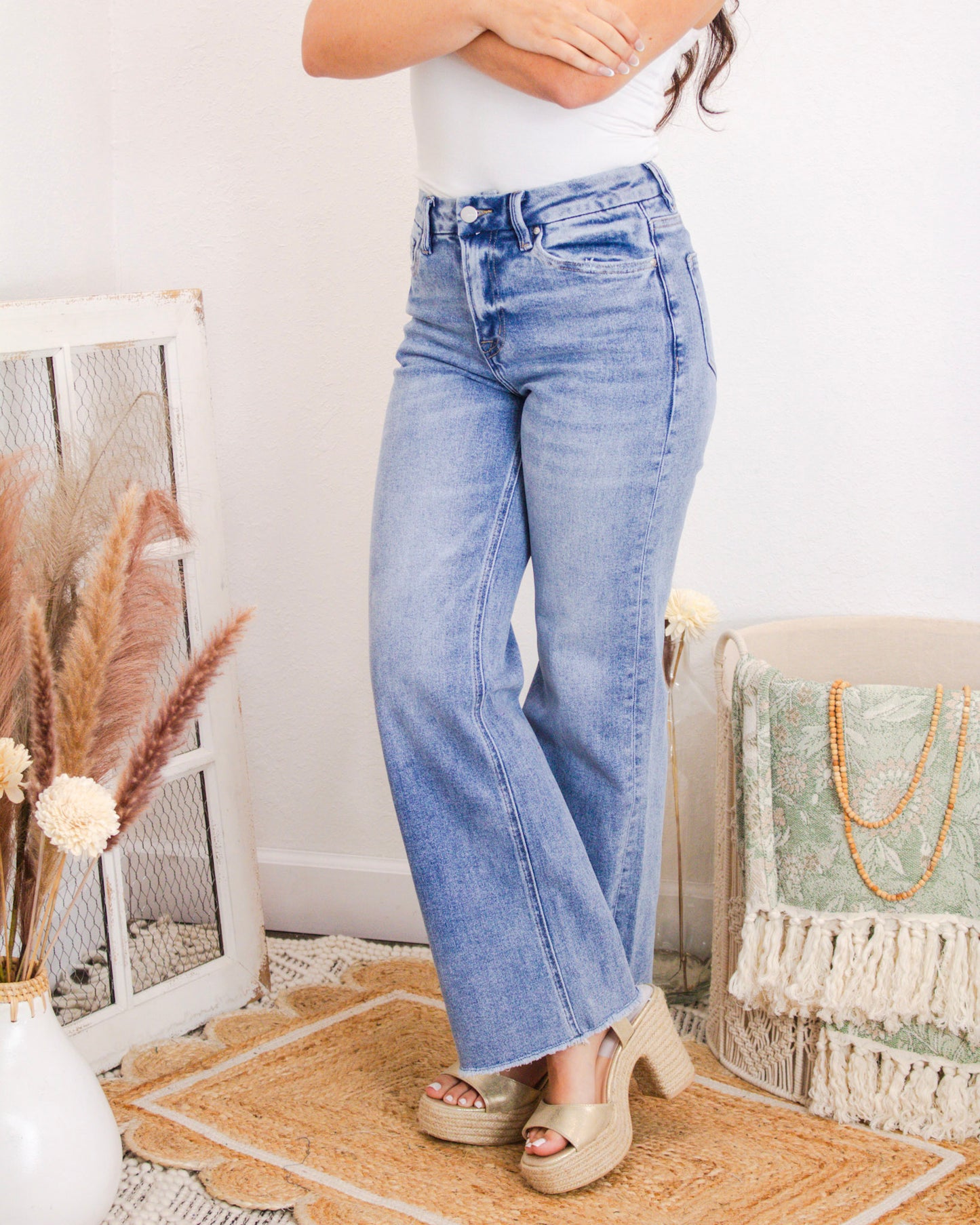 Front view of a woman wearing high-rise light wash jeans with a subtle flare and raw hem, paired with a white tank top and platform sandals.