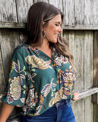 Woman wearing a dark teal floral print blouse with wide sleeves and button details, styled with dark denim jeans for a relaxed yet chic boutique look.
