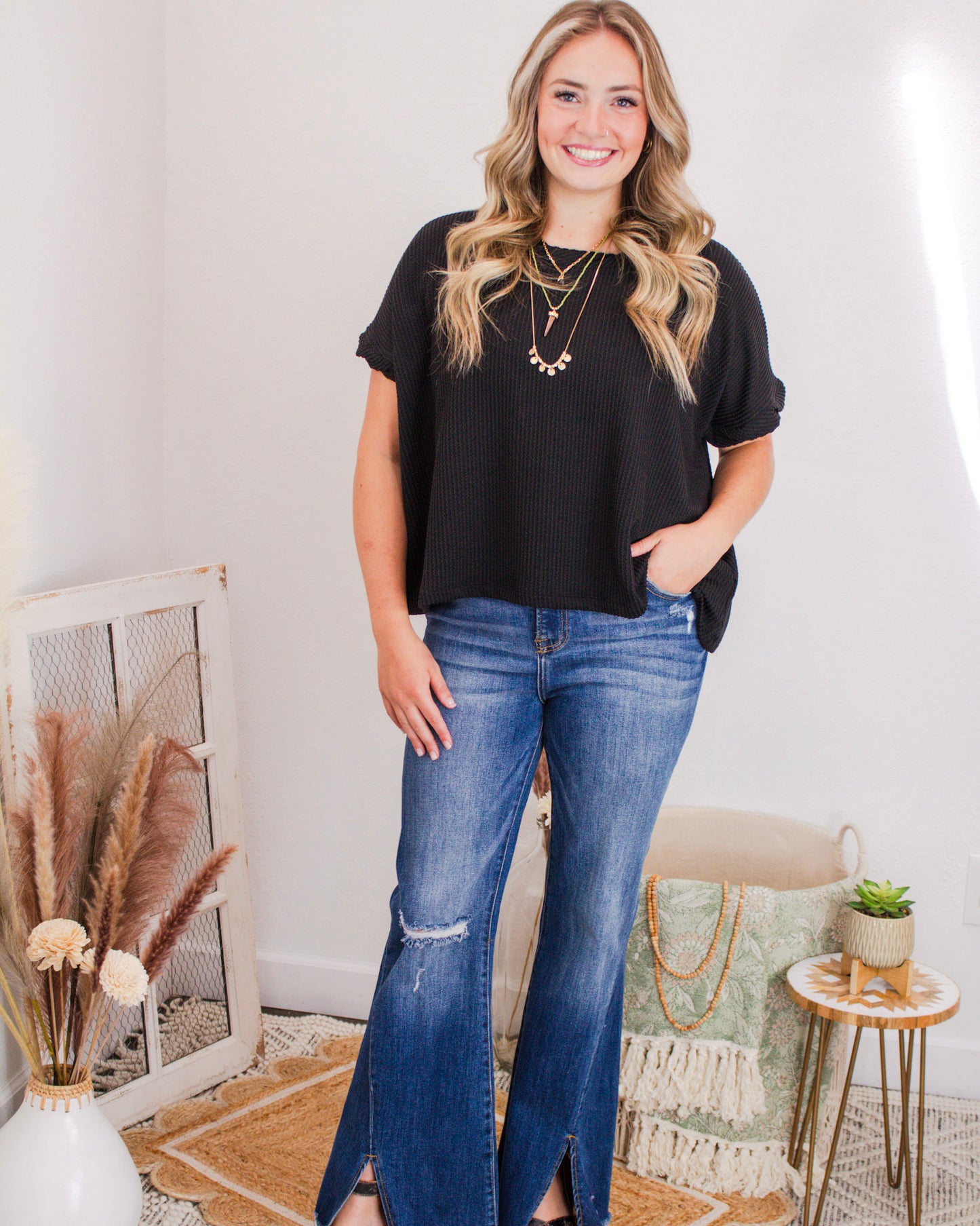 Woman modeling a black textured short-sleeve top with layered necklaces and flared slit-hem jeans in a modern boho-style room.