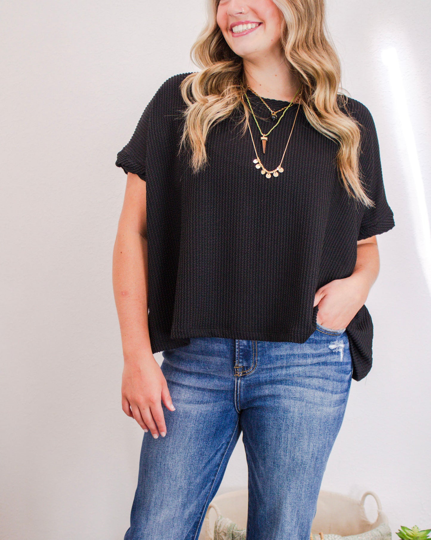 Woman wearing a black textured short-sleeve top with layered gold necklaces and mid-wash jeans.