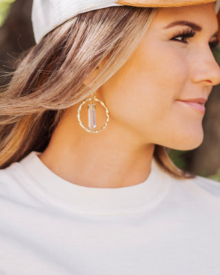 Smiling woman wearing a gold hoop earring with a clear quartz crystal pendant, styled with a white top and trucker hat for a chic casual look.
