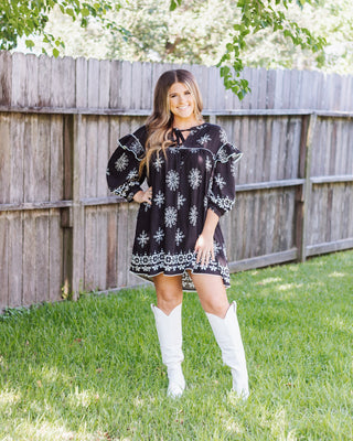 Black dress with white embroidered floral patterns and ruffled details, styled with high white cowboy boots