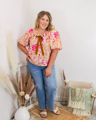Woman modeling floral puff sleeve top with mustard bow tie detail, paired with straight leg jeans and sandals—boho-chic style at Makenzie Layne Boutique