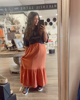 Woman taking a boutique mirror selfie in a black graphic tee and burnt orange maxi skirt with ruffle hem, styled with platform sandals.