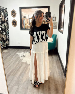 Woman taking a mirror selfie in a boutique, wearing a black TEXAS graphic tee, white tiered maxi skirt with front slit, and black embellished sandals.