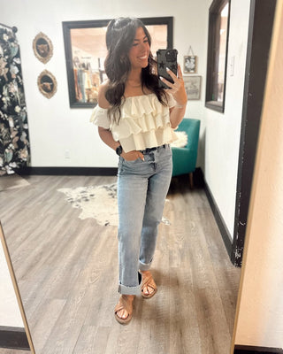 Woman taking a mirror selfie in a boutique, wearing an off-the-shoulder cream ruffle top, light-wash straight leg jeans with a cuffed hem, and tan criss-cross sandals.