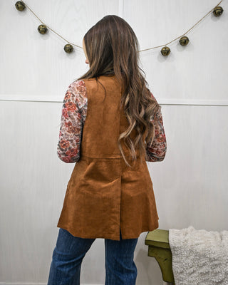 Woman wearing a brown suede vest over a floral shirt and jeans, standing against a white wall with string lights.