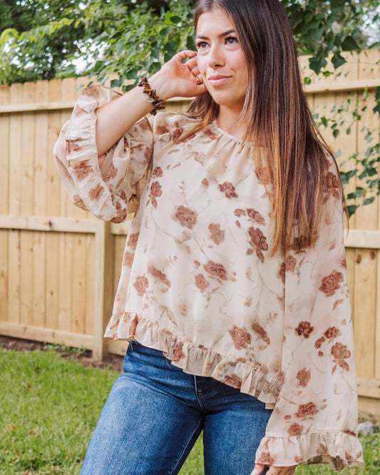 Woman wearing a beige floral long sleeve blouse with ruffle hem and bell sleeves, paired with blue jeans for a feminine and casual chic look.