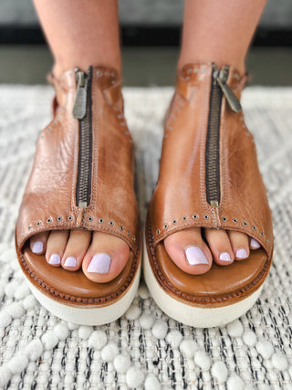 Close-up of brown leather platform sandals with front zipper, stud detailing, and white sole on woven rug