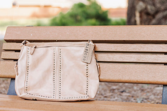 Beige Bed Stu leather crossbody bag with brass stud detailing, displayed on a wooden bench. Stylish handcrafted bag for everyday wear or boho-chic outfits.
