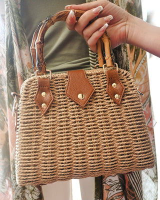 Close-up of a woven straw handbag with bamboo top handles, vegan leather accents, gold hardware, and a detachable crossbody strap—perfect for summer, vacation, and resort style.