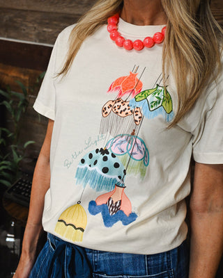 Women's ivory graphic tee featuring colorful vintage-inspired umbrellas and “Be the Light” design, styled with a bold coral statement necklace for a playful boutique look.