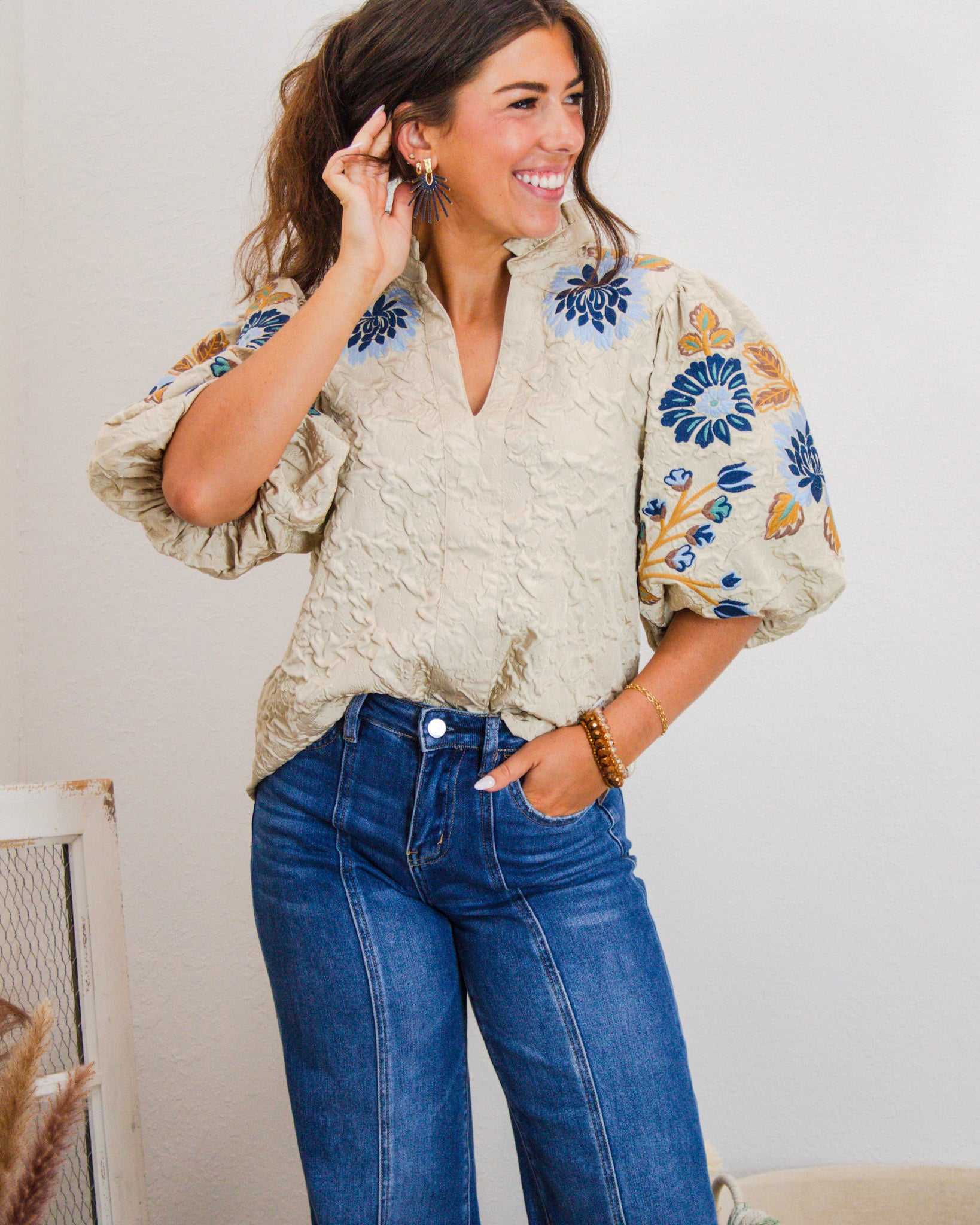 Woman wearing a beige embroidered puff sleeve blouse with blue and mustard floral details, paired with high-rise wide-leg jeans.