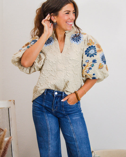 Woman wearing a beige embroidered puff sleeve blouse with blue and mustard floral details, paired with high-rise wide-leg jeans.