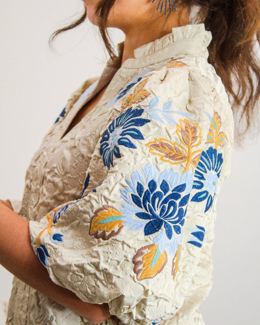 Close-up of a beige textured blouse with puff sleeves featuring blue, mustard, and brown floral embroidery.