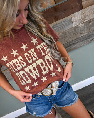 Women’s rust graphic tee with “Dibs on the Cowboy” vintage western print, styled with distressed denim shorts and a statement belt for a trendy boutique look