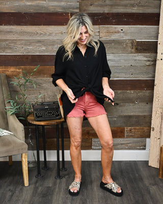 Women's black oversized button-down blouse styled with coral frayed hem shorts and platform sandals, creating a chic casual boutique outfit perfect for summer, vacations, and everyday wear.