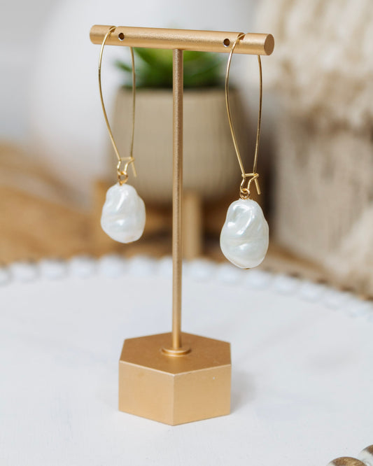 Pair of modern gold teardrop hoop earrings with abstract white pearl-like drops displayed on a gold stand against a neutral background with natural decor.
