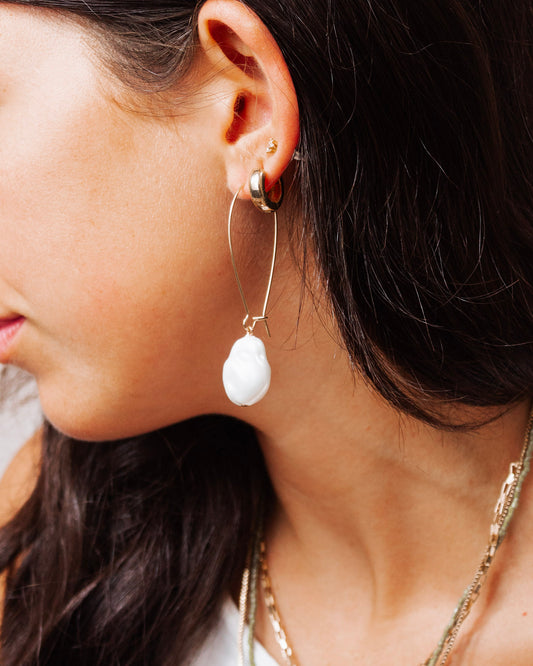 Close-up of a woman's ear wearing a modern gold hoop earring with a large white abstract bead drop, styled with layered gold necklaces and dark hair.