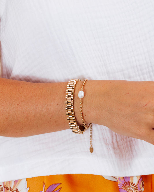 Woman wearing a stylish gold bracelet stack with a pearl-beaded bracelet and link band, paired with a white top and orange floral skirt.