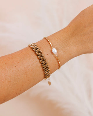 Close-up of a woman's wrist wearing a gold bracelet stack with a wide link band and a delicate pearl-beaded bracelet, styled against a soft neutral background.