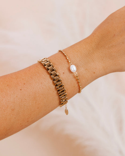 Close-up of a woman's wrist wearing a gold bracelet stack with a wide link band and a delicate pearl-beaded bracelet, styled against a soft neutral background.