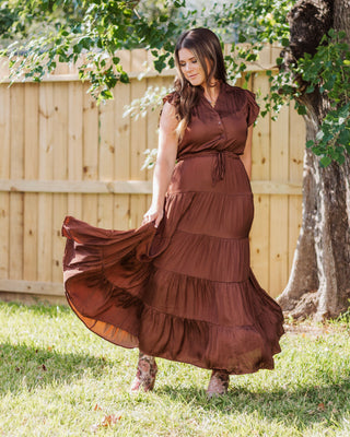 Woman wearing a flowing chocolate brown tiered maxi dress with a drawstring waist, styled outdoors with ankle boots for an elegant fall look.