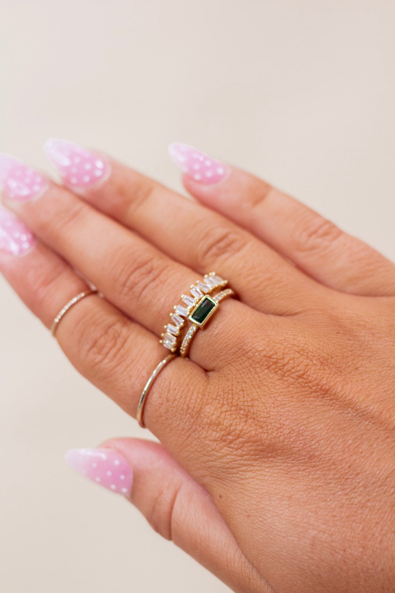 Close-up of hand wearing a set of gold stackable rings featuring a rectangular green gemstone centerpiece, baguette-cut clear crystals, and pavé accents. Elegant and modern ring set perfect for special occasions or everyday glam.