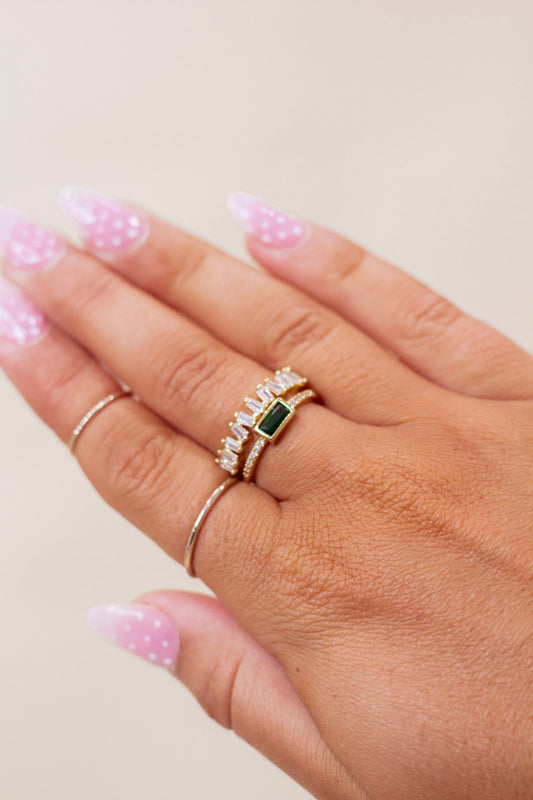 Close-up of hand wearing a set of gold stackable rings featuring a rectangular green gemstone centerpiece, baguette-cut clear crystals, and pavé accents. Elegant and modern ring set perfect for special occasions or everyday glam.