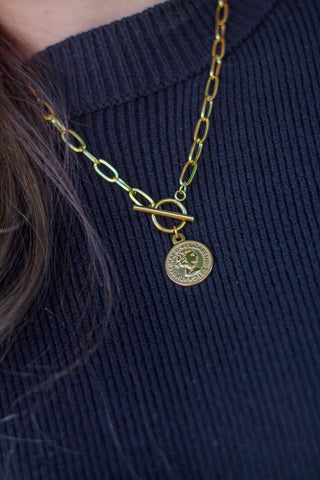 Close-up of a gold coin pendant necklace with toggle clasp on a chunky chain, styled over a ribbed black knit sweater for a timeless, elegant look.