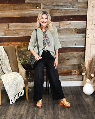 Woman wearing a sage green knit top with black wide leg jeans, patterned neck scarf and crossbody bag for a chic casual boutique outfit.