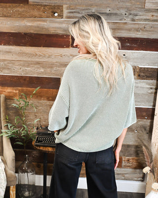 Women's sage green oversized knit sweater with a relaxed draped fit, short sleeves and textured ribbed design, styled with black denim pants.