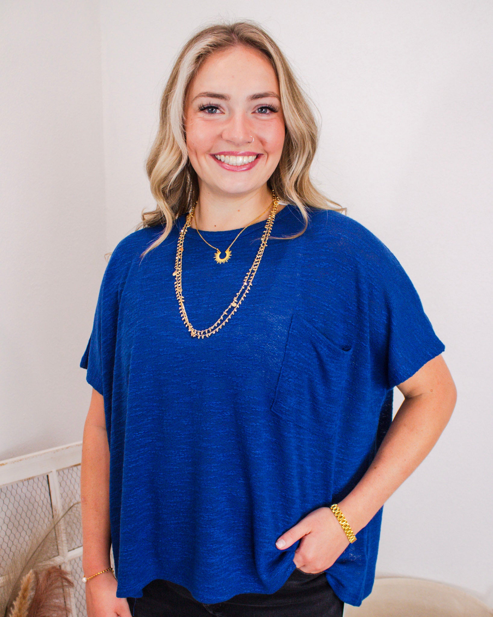 Smiling woman wearing a cobalt blue short-sleeve top with a front pocket, layered gold necklaces, and a gold bracelet.