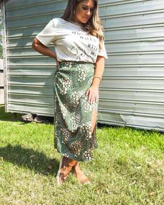 Woman styling a “Give Me Jesus” faith tee tucked into a green floral midi skirt with side slit, paired with tan sneakers for a chic, casual boutique look.
