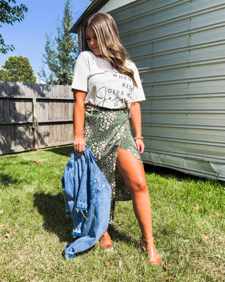 Woman wearing a “Give Me Jesus” graphic tee tucked into a green floral slit skirt, holding a denim jacket for a chic and faith-inspired boutique outfit.