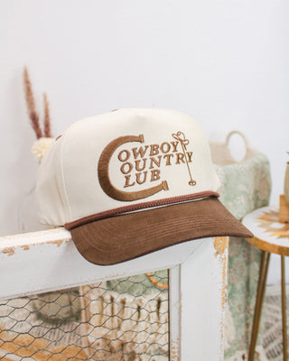 Cream and brown Sendero Provisions Co. “Cowboy Country Club” hat featuring embroidered western golf logo, displayed on a rustic wooden chair indoors.