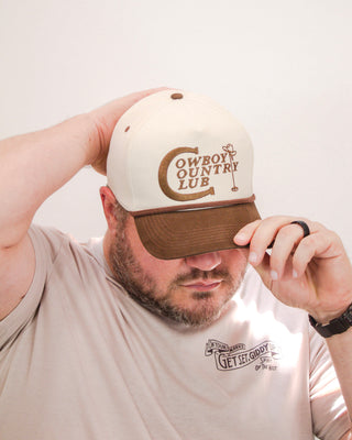 Man wearing a cream and brown Sendero Provisions Co. hat with “Cowboy Country Club” embroidered logo, styled with a western graphic tee and casual pose.
