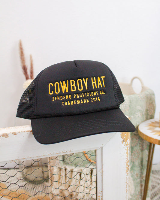 Black mesh trucker hat with bold yellow embroidered text reading “Cowboy Hat – Sendero Provisions Co. Trademark 2014,” displayed on a rustic white wood frame.