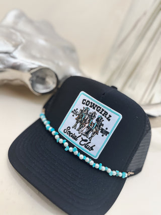 Black trucker hat with a "Cowgirl Social Club" patch featuring illustrated cowgirls, accented with a turquoise and pearl hat band, styled beside a silver cow skull.