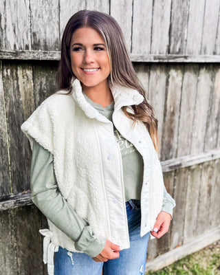 Woman wearing a soft white sherpa vest layered over a sage green long sleeve top and distressed jeans, styled for a cozy, casual fall outfit.