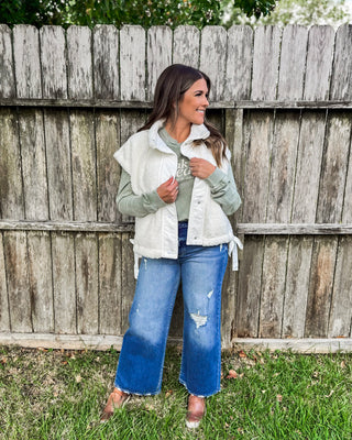 Woman wearing a cozy white sherpa vest over a sage green sweatshirt with wide-leg distressed jeans and brown shoes, styled for a casual fall outfit.