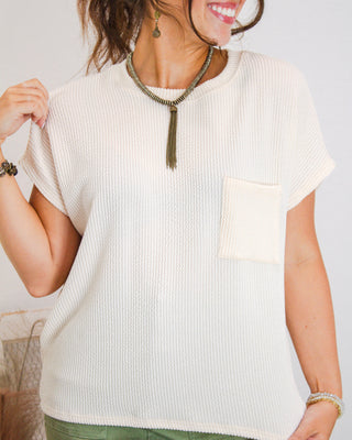 Close-up of a woman wearing a cream textured short sleeve pocket top with olive accessories, styled for a chic casual look from MaKenzie Layne Boutique.