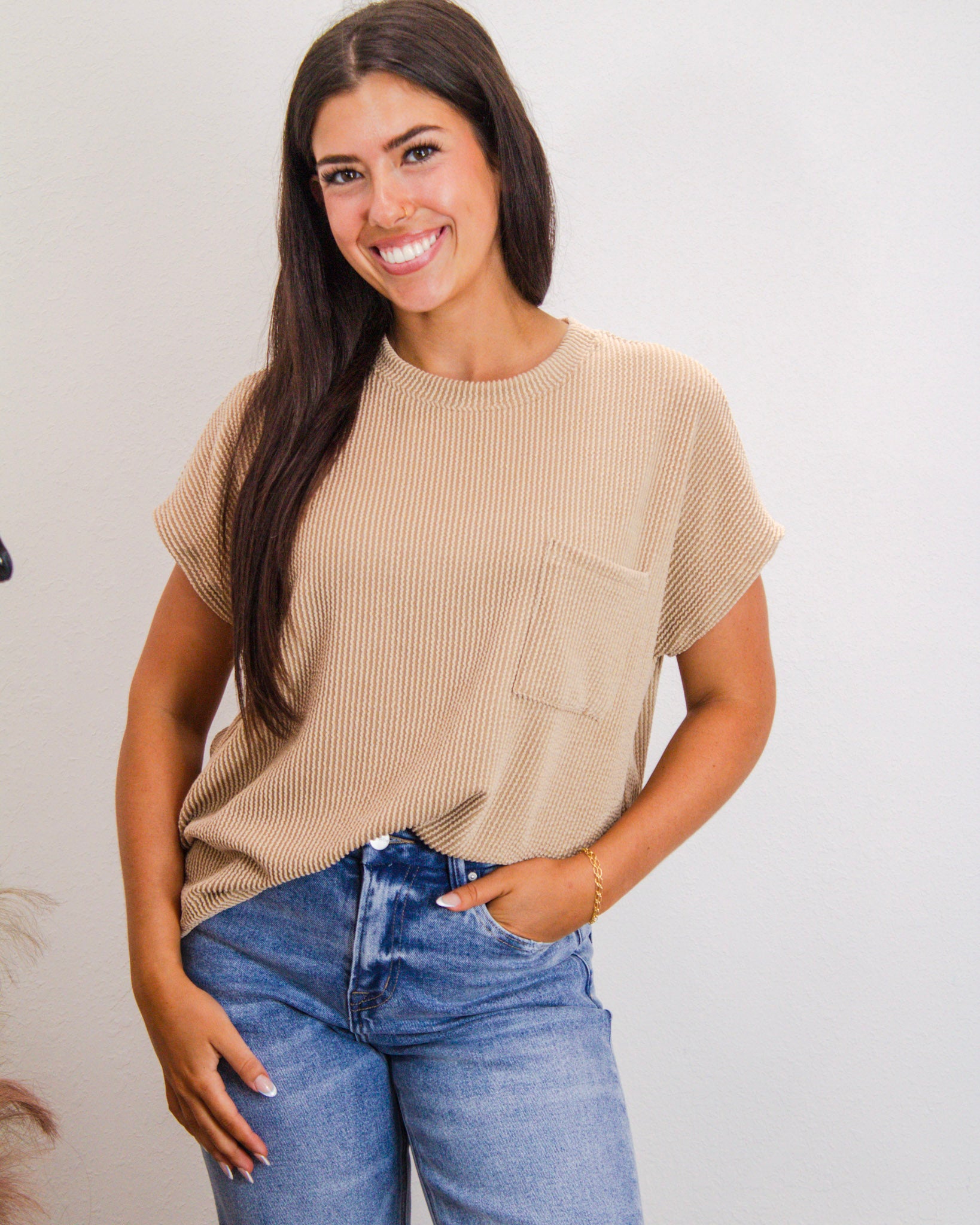 Woman wearing a beige ribbed short sleeve knit top with front pocket, paired with high-rise blue jeans, styled for a casual boutique look.